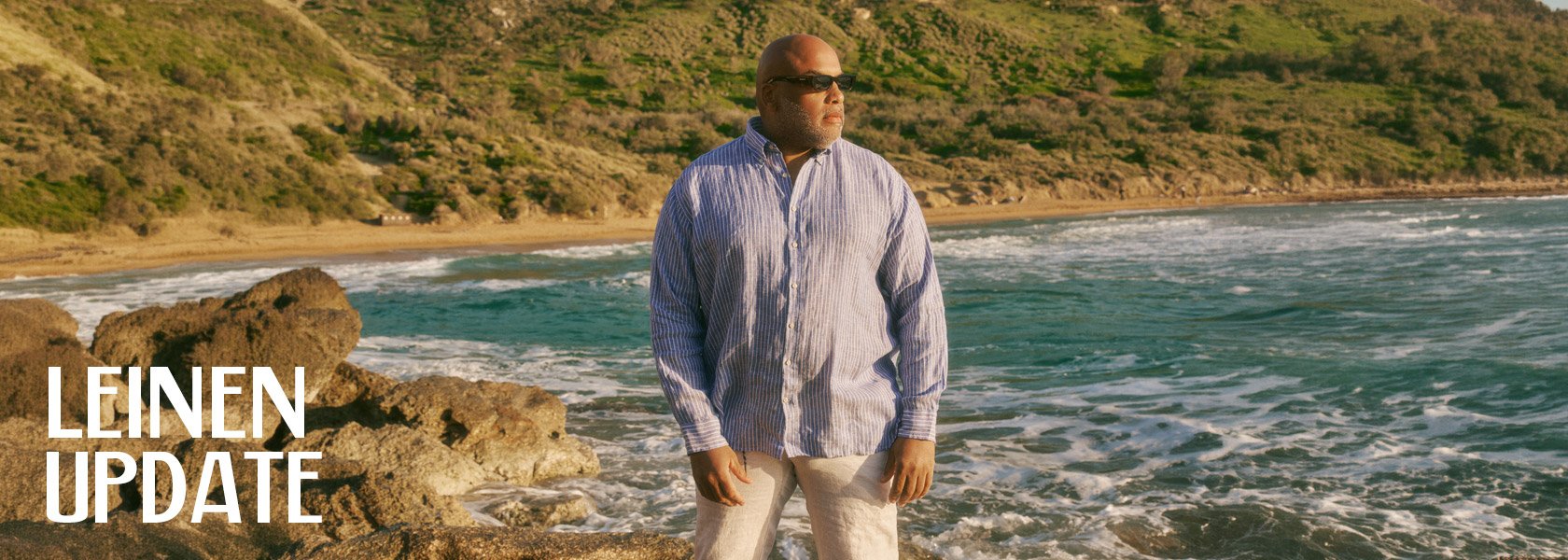 Ein Mann in einem gestreiften Hemd und hellen Hosen steht an einem felsigen Strand mit Wellen und sanften Hügeln im Hintergrund.