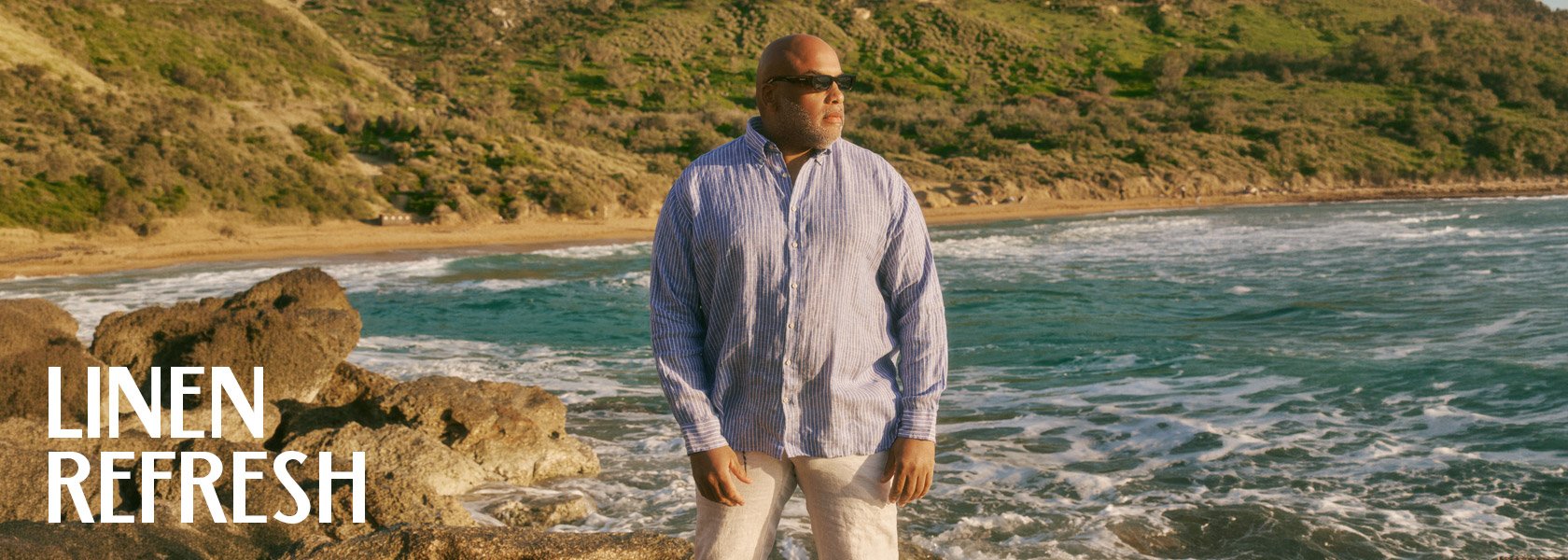 A man wearing a striped linen shirt and light pants stands on rocks by the ocean.