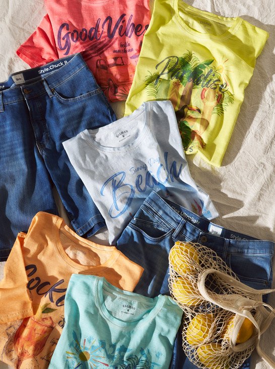 Mehrere bunte T-Shirts mit Schriftzug und Aufdrucken liegen neben Jeans und einer Netztasche mit Zitronen.