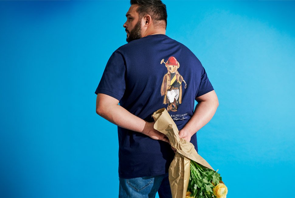 A man, seen from the back, wearing a navy blue t-shirt with a Ralph Lauren Polo Bear design, holding a bouquet wrapped in brown paper. The background is bright blue. He's wearing blue jeans.