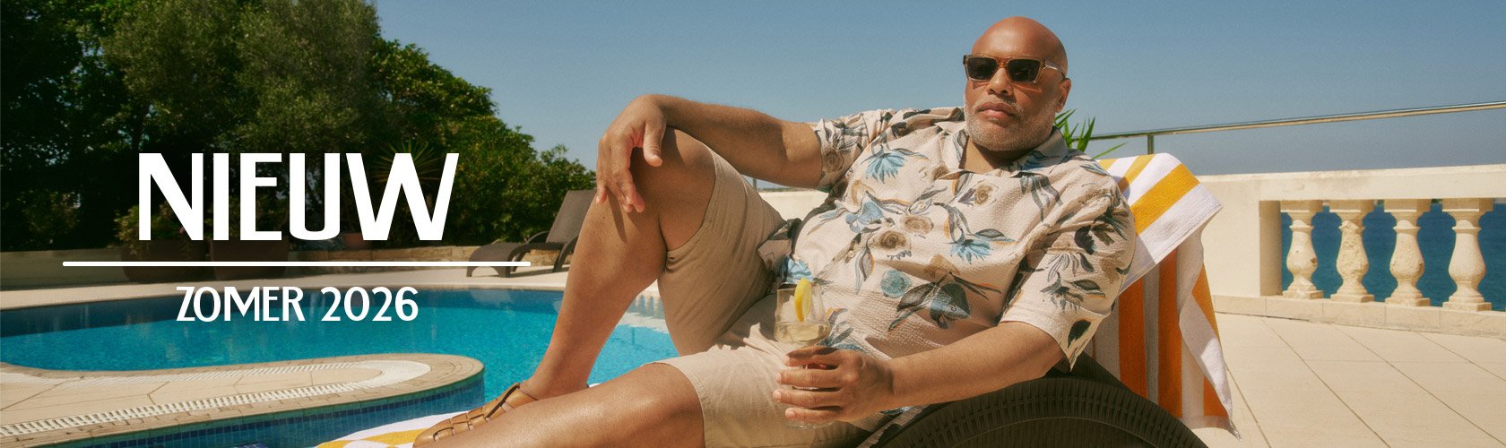 Man in een zomerse blouse en korte broek ontspant bij een zwembad met een drankje.