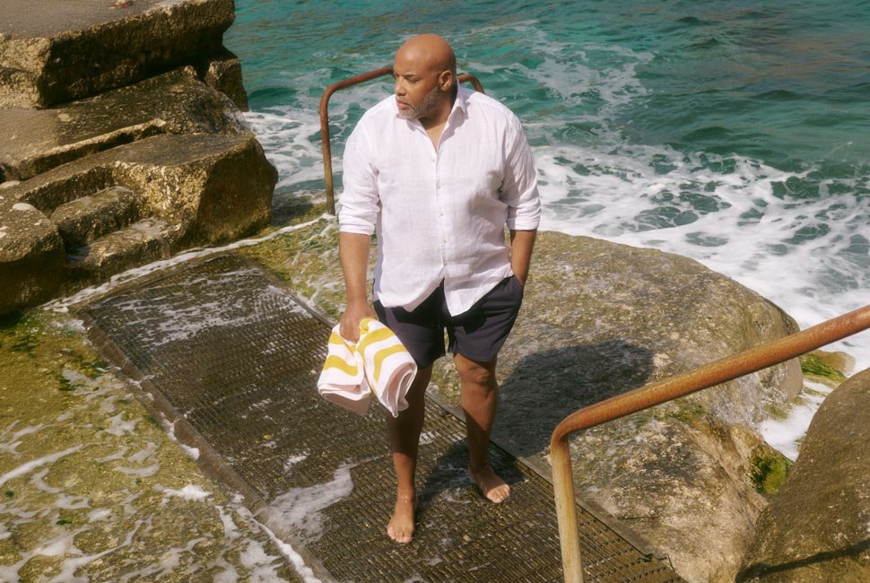 A man in a white shirt and dark shorts stands on wet rocks by the sea, holding a striped towel.