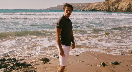 Ein Mann in einem dunklen Poloshirt und weißen Shorts steht am Strand mit Wellen und Felsen im Hintergrund.