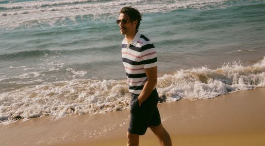 Mann mit Sonnenbrille und gestreiftem Poloshirt geht am Strand entlang, die Wellen brechen am Ufer