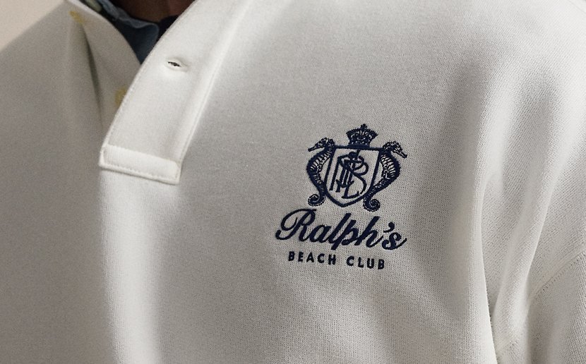 Na einem weißen Poloshirt ist das Emblem von Ralphs Beach Club in dunkelblau aufgestickt.
