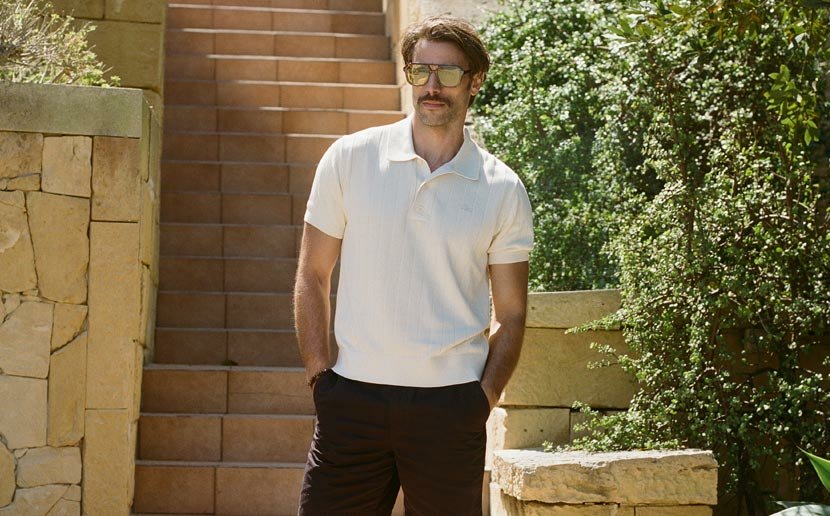 Mann mit Schnurrbart trägt cremefarbenes Poloshirt und dunkle Shorts, steht neben einer Steintreppe und üppiger grüner Vegetation.