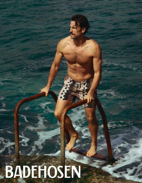 Ein Mann mit Schnurrbart und Badeshorts hält sich an einer rostigen Leiter fest, während er ins Meer steigt