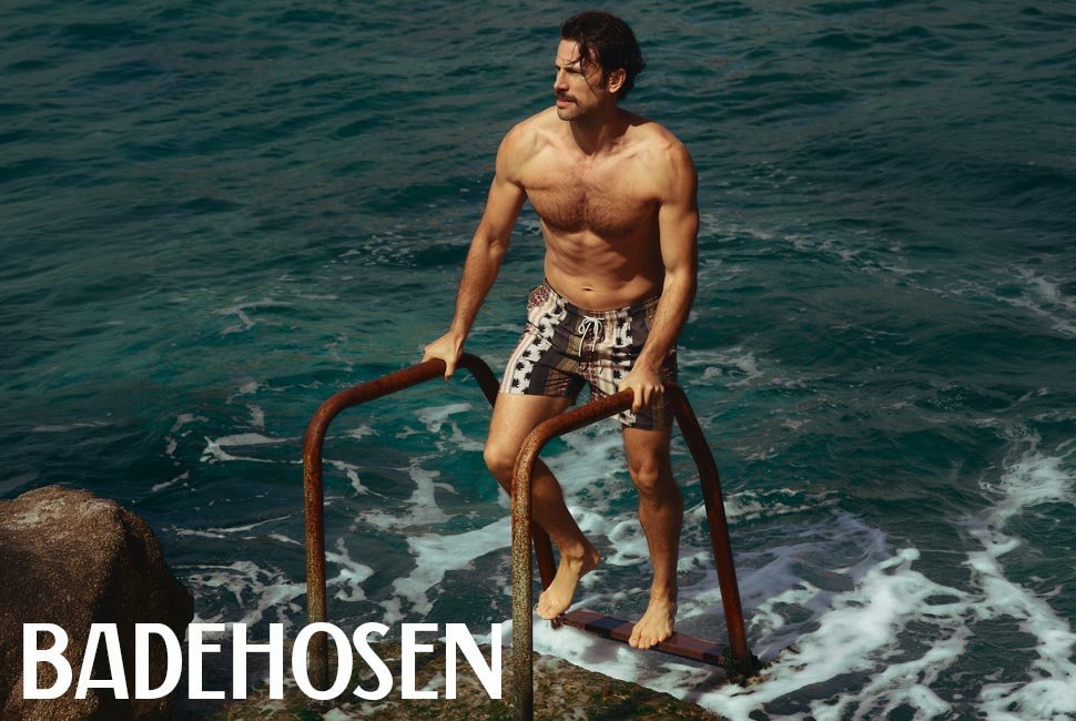 Ein Mann in Badeshorts steigt über eine rostige Leiter ins türkisfarbene Meer.