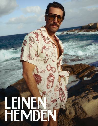 Ein Mann mit Sonnenbrille und Schnurrbart trägt ein kurzärmeliges Leinenhemd mit Muster am Meer.