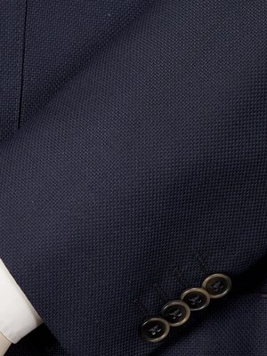 Close-up view of navy blue suit fabric with a textured weave. The image shows a portion of a suit sleeve, including four dark buttons. A small amount of white shirt cuff is visible.