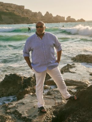 Ein Mann mit Sonnenbrille und gestreiftem Hemd steht an einem felsigen Strand bei Sonnenuntergang.
