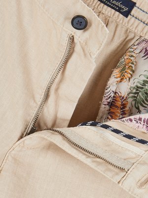 Nahaufnahme beiger Herrenhose mit Reißverschluss und Knopf.  Das Innenfutter zeigt ein tropisches Muster.  Ein Label ist sichtbar.