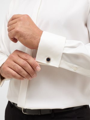 Nahaufnahme eines Mannes, der Manschettenknöpfe an einem weißen Hemd anlegt. Der Mann trägt ein weißes Hemd mit verdeckter Knopfleiste. Zu sehen sind auch ein schwarzer Gürtel und Hosen. Der Manschettenknopf hat einen dunklen Kreis.
