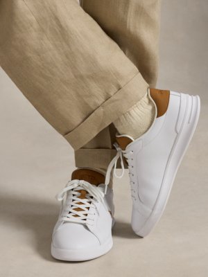 Nahaufnahme der Beine einer Person, die beigefarbene Hose und weiße Turnschuhe trägt. Die Turnschuhe sind aus Leder mit braunen Details und Schnürsenkeln. Der Hintergrund ist hell. Die Hose ist bis zum Knöchel hochgekrempelt.