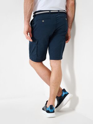 A man stands facing a white wall wearing navy cargo shorts, a white t-shirt, and a belt. He wears black and blue sneakers. The photo is taken from the rear, showing the back and side.
