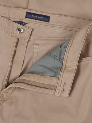 Detailní záběr na světlé kalhoty, které jsou zčásti rozepnuté. Viditelný je zip, zapínání na knoflík a štítek Pierre Cardin. V rozepnuté části je vidět světle modrá podšívka.