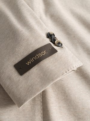 Nahaufnahme des Ärmels eines beigefarbenen Blazers. Auf dem Ärmel sind drei Knöpfe zu sehen. Ein Etikett mit dem Wort "windsor." ist ebenfalls angenäht. Das Material des Blazers scheint grob gewebt zu sein.