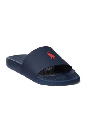Ein blauer Polo-Slipper, der auf einer weißen Oberfläche liegt. Auf dem Riemen des Schuhs ist ein rotes Logo mit einem Reiter zu sehen. Der Slipper hat eine geriffelte Sohle.