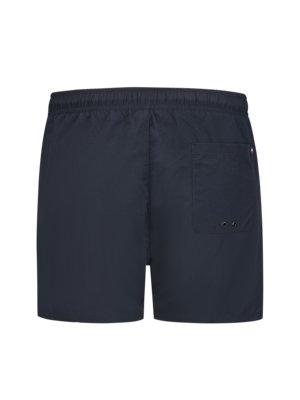 Dunkelblaue Badeshorts mit einem Gummibund. Auf der Rückseite befindet sich eine aufgesetzte Tasche und ein kleines Logo. Zwei kleine Löcher sind an der Seite der Tasche.