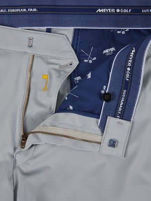 Nahaufnahme einer geöffneten, hellgrauen Golfhose. Der Reißverschluss ist geöffnet. Innenfutter ist dunkelblau mit weißen Golfschläger-Grafiken und Logo. Markenschriftzüge sind sichtbar. Ein Knopf und ein kleiner Knopfverschluss sind erkennbar.