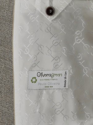 Nahaufnahme der Innenseite eines Anzugs. Ein brauner Knopf an einem weißen Stoff. Der Stoff hat ein geprägtes Muster. Ein weißes Etikett mit dem Schriftzug "Oliveiragreen", einem Recycling-Symbol und "Paulo Oliveira" ist aufgenäht.