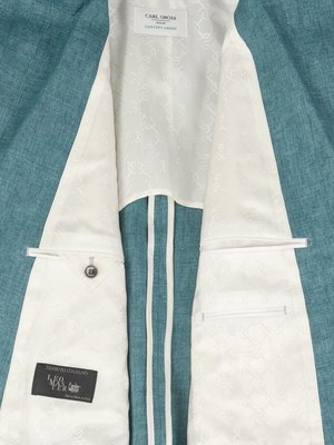 Nahaufnahme eines blauen Sakkos, dessen Innenseite zu sehen ist. Das Futter ist weiß mit einem Muster. Links ist eine Tasche mit einem Knopf. Auf der Innenseite ist ein Etikett mit Schrift. Rechts eine Tasche. Oben ein Label mit Schrift.