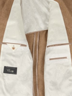 Detailní pohled na vnitřek hnědého saka. Bílá podšívka s jemným vzorem zdobí vnitřní stranu. Dvě kapsy s lemováním, jedna s knoflíkem. Štítek "LEO MAS PER" dole vlevo.
