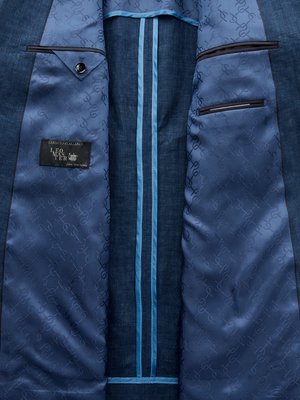 Close-up of the inside of a blue suit jacket. The jacket's interior is lined with patterned blue fabric. Two pockets, one with a button, are visible, and there's a tag with text. Two light blue stripes run vertically down the jacket's center.