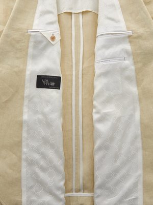 Ansicht von innen einer hellbraunen Jacke. Das Innenfutter ist weiß mit einem Muster aus sich wiederholenden Symbolen. Auf der linken Seite ist ein kleiner Knopf und ein Etikett mit schwarzem Hintergrund sichtbar. Die Jacke hat zwei Innentaschen.