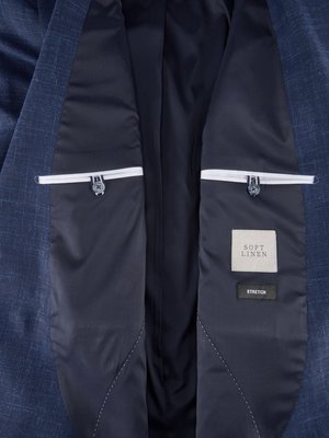 Detailansicht eines blauen Sakkos, innen offen. Links und rechts jeweils eine Brusttasche mit Knopf. Auf der rechten Seite Etiketten mit der Aufschrift "SOFT LINEN" und "STRETCH". Das Futter ist dunkelblau.
