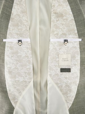 Nahaufnahme des Innenfutters eines hellgrauen Sakkos. Beide Innenseiten sind mit cremefarbenem, floral gemustertem Stoff ausgekleidet. Weiße Stoffhalter mit Knöpfen, ein Label "Soft Linen" und eine schwarze Textilmarke sind zu sehen.
