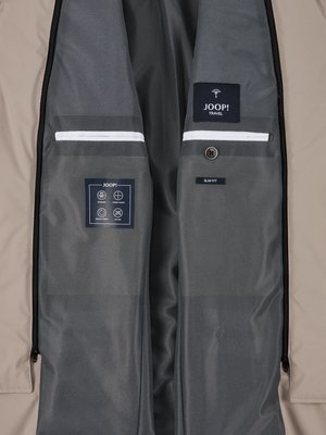 Nahaufnahme des geöffneten Innenraums einer beigefarbenen Jacke. Man sieht zwei Innentaschen mit weißen Paspeln. Auf einer Tasche befindet sich ein JOOP!-Label, auf der anderen ein JOOP! Travel-Label. Ein graues Innenfutter ist sichtbar.