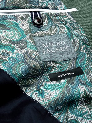 Nahaufnahme des Innenteils einer grünen Jacke mit Paisley-Muster. Ein graues Etikett mit der Aufschrift "MICRO JACKET" und ein schwarzes Etikett mit "STRETCH". Oben ein Knopf an einem blau-weiß gestreiften Stoff.