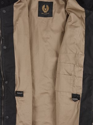 Interior view of a black Belstaff jacket. The jacket is open, revealing a tan lining with a pocket. A black label with gold lettering is visible near the top. Metal hardware is visible.