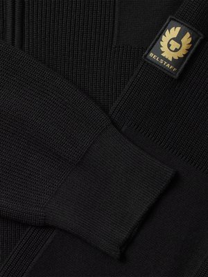 Close-up shot of black knitwear. The image shows the arm and part of the body of a sweater. A gold and black Belstaff logo is on the sleeve. The knitwear has a ribbed texture.