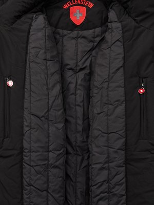 Nahaufnahme einer schwarzen Jacke, geöffnet, mit gestepptem Innenfutter. Oben das Wellensteyn-Logo mit rotem Schild und Kreuz. Zwei Reißverschlusstaschen mit Anhängern auf der Jacke.