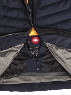 Detailaufnahme einer dunkelblauen Steppjacke. Die Jacke ist teilweise geöffnet und zeigt das Innenfutter. Auf dem Brustbereich ist ein rotes Emblem mit weißem Kreuz erkennbar. Der Reißverschluss verläuft zentral.