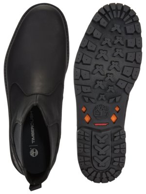 A top-down view of a black Timberland Chelsea boot and the boot's outsole. The boot has a leather upper and the sole is textured with the Timberland tree logo and orange diamond shapes. The other shoe's sole is also visible.