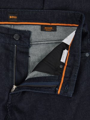Nahaufnahme einer Jeanshose mit offenem Reißverschluss. Auf dem Stoff sind die Marken BOSS und REMAIN sichtbar. Der Hosenbund hat einen orangefarbenen Streifen. Der Reißverschluss ist metallfarben. Ein Knopf mit dem Logo der Marke ist sichtbar.