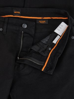 Nahaufnahme einer schwarzen Jeanshose mit geöffnetem Reißverschluss. Auf dem Bund sind die Logos "BOSS" und "REMAINE" zu sehen. Im Innenteil der Hose sind Labels angebracht. Der Saum des Innenfutters ist mit orangefarbenem Stoff verziert.