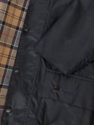 Nahaufnahme einer dunkelblauen Jacke mit Knopfleiste und einer braunen Innenseite mit Karomuster. Das Etikett "Barbour" ist auf der Brusttasche zu sehen. Die Jacke hat einen Kragen und Ärmel.