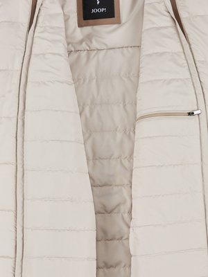 Nahaufnahme einer hellbeige gesteppten Jacke, geöffnet. Ein Reißverschluss verläuft entlang der Jackenkante. Das Markenlogo "JOOP!" ist am oberen Rand zu sehen. Innenfutter und Außenseite der Jacke sind in hellen Farbtönen gehalten.