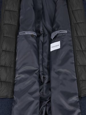The inside of a dark blue, quilted, long winter coat. Gray fabric lines the pockets and collar. A white tag that reads "TOM RUSBORD" is visible at the bottom of the coat. The coat is open.
