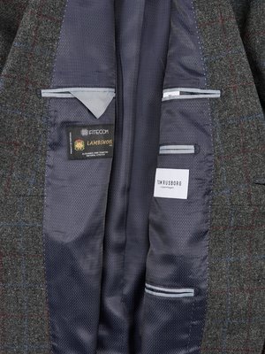 Detailní záběr vnitřku pánského saka v šedé barvě s modro-červenou kostkou. Jsou viditelné kapsy a štítky s nápisy FITECOM LAMBSWOOL a TOM RUSBORG Copenhagen. Materiál vnitřní podšívky je tmavě šedý s jemným vzorem.