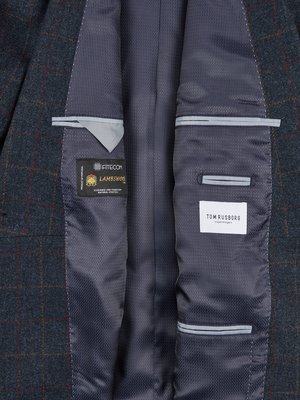 Detail uvnitř tmavě modrého saka s kostkovaným vzorem. Jsou viditelné dvě vnitřní kapsy a štítky s nápisy "FITECOM LAMBSWOOL" a "TOM RUSBORG".  Látka má jemnou texturu a lemování.