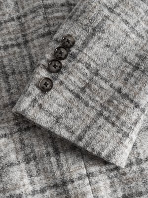 Nahaufnahme eines grauen Blazerärmels aus Tweed mit Knöpfen. Der Stoff hat ein dezentes Karomuster. Vier Knöpfe sind entlang des Ärmels aufgenäht. Die Knöpfe sind dunkelgrau und rund.