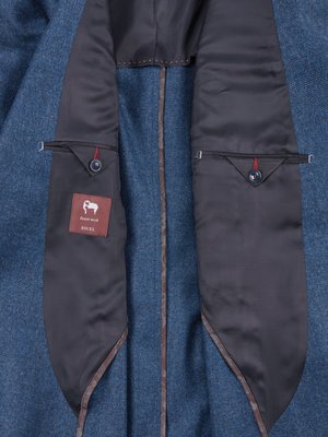 Innenansicht eines blauen Anzugs. Geöffnet, Innenseite sichtbar. Dunkelgraues Innenfutter, zwei Taschen mit Knöpfen. Eine Tasche mit einem Etikett, das ein Schaf und Text zeigt.