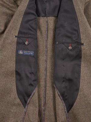 Close-up view of the interior of a brown blazer. Two black satin-lined pockets are visible, each with a button and a small rectangular flap. A label is sewn into the left pocket. The blazer's lining is visible.