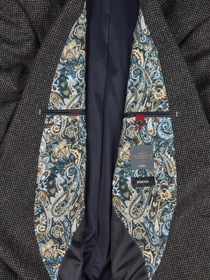 Nahaufnahme eines dunkelgrauen Sakkos. Innenfutter mit Paisleymuster in Blau-, Beige- und Grüntönen. Die Innentaschen sind mit rotem Knopf und schwarzer Paspel abgesetzt. Ein Etikett mit Schriftzug ist sichtbar.