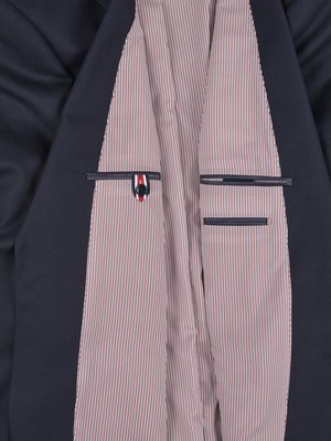 Nahaufnahme des Innenteils eines dunkelblauen Sakkos. Innenfutter mit rot-weiß gestreiften Stoff, rechts und links jeweils eine Innentasche mit schwarzer Paspelierung. Auf einer Innentasche ist ein Emblem in rot-weiß-blau zu sehen.